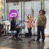 A keyboard player, violinist and vocalist perform a song in a small performance space surrounded by glass windows in downtown Boston.