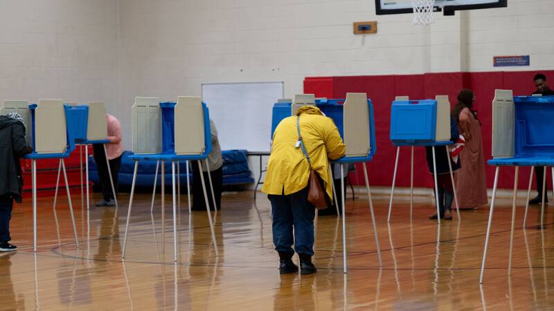 Several people fill out their ballots in private desks in what looks like a high school gym.