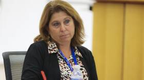A woman sits at a conference room table, holding a pen in her right hand poised to take notes while she listens to a speaker.
