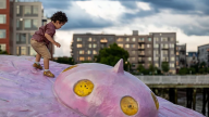 A young child leans over to maintain their balance while standing atop a slanted, pink UFO sculpture. Large city buildings are in the background.