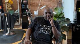 A man smiles as he sits in a chair at a barbershop, one hand resting against his face.