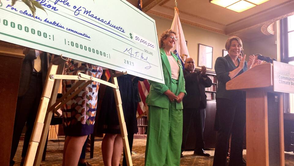 A large green check for $50 million, made out to "The people of Massachusetts" stands on an easel next to Gov. Maura Healey, who stands behind a lectern clapping.