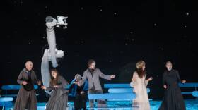 This is a photo from the play The Orchard. It is winter. The stage is bathed in blue light.  Six people are outside in what appears to be a park filled with park benches. They are laughing and look joyous. In the background is a large robotic arm that looks like a huge telescope