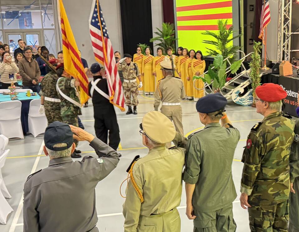 A row of elderly men in military uniforms salute as a color guard marches past bearing the flags of the United States and South Vietnam during an indoor ceremony of the 50th anniversary of the end of the Vietnam War.