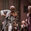A man in a powdered wig and formal gray coat plays a violin.