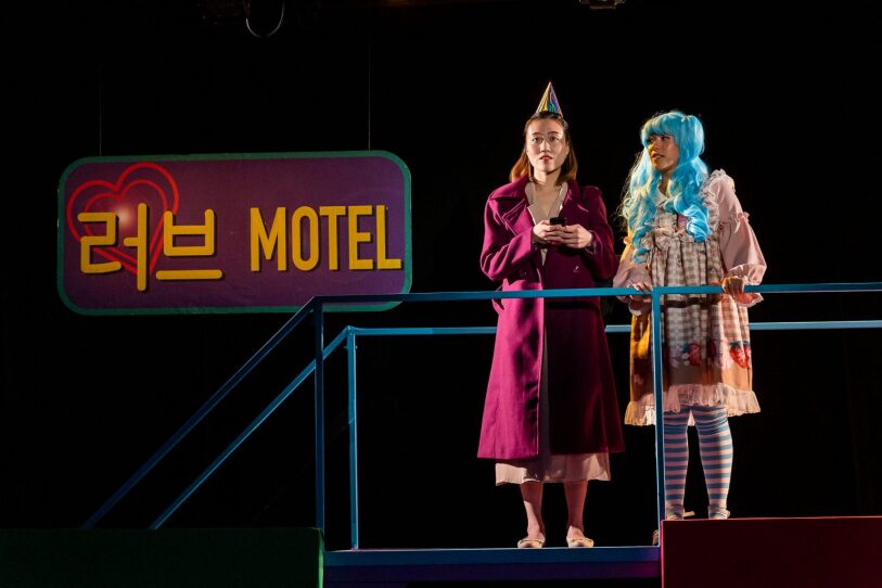 two actors, one in a party hat and one in a blue wig, stand at a railing in front of a sign in a production of "Wild Goose Dreams"