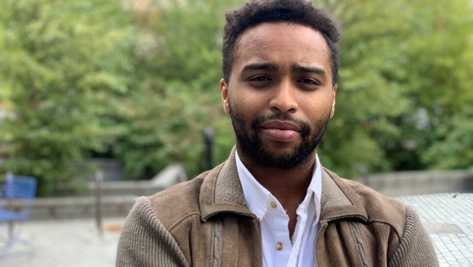 A young man with a worn expression on his face stands in a courtyard.