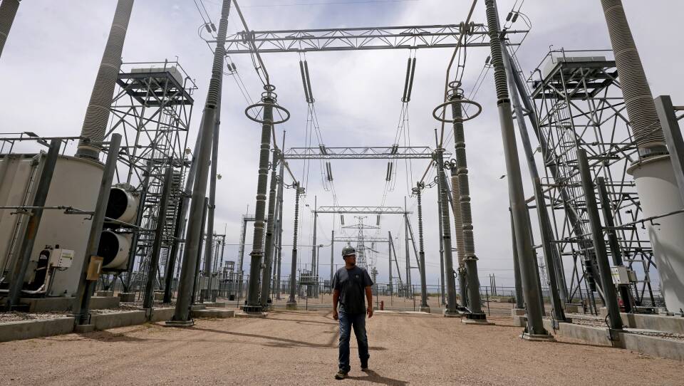 A worker in a hard hat walks on the dirt ground in between large electricity towers.