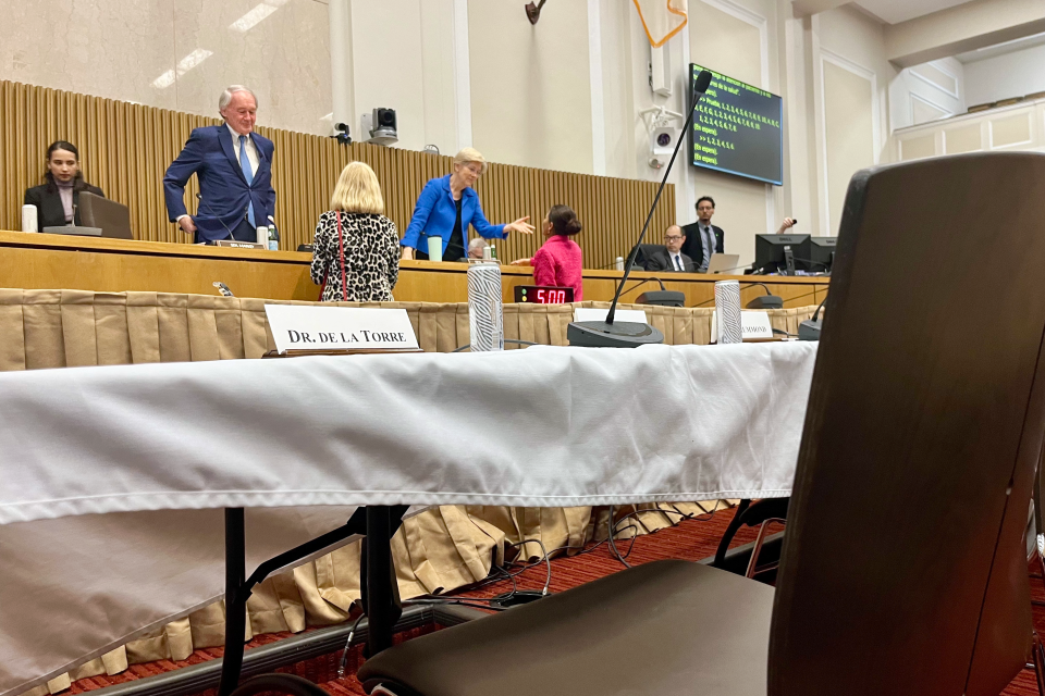 A placard with the words "Dr. De La Torre" sits on a table covered with a white cloth, with an empty chair in front of it. Senators Ed Markey and Elizabeth Warren are visible in the background.