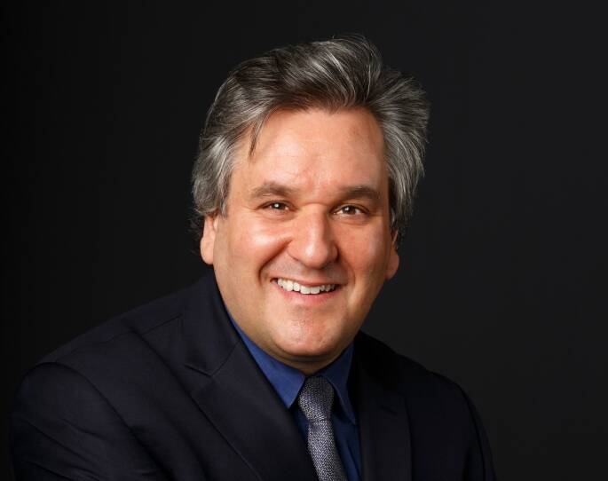 A man wearing a suit smiles into the camera in a staged headshot on a dark background