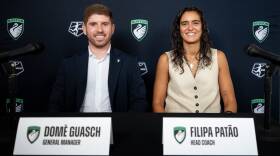 A man in a blue jacket and white shirt sits next to a woman in a tan shirt. Both are in front of a black backdrop and have their names and titles on name cards listed in front of them.