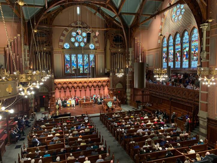 People fill the pews inside a church, including a second level underneath stained glass windows.