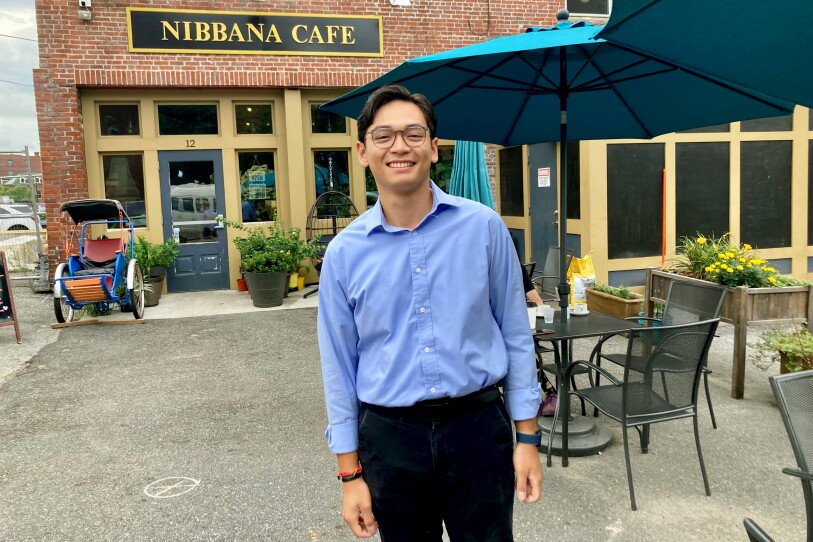 A man with black hair and glasses stands on the patio of a coffee shop, with a brick building and umbrella-covered table behind him.