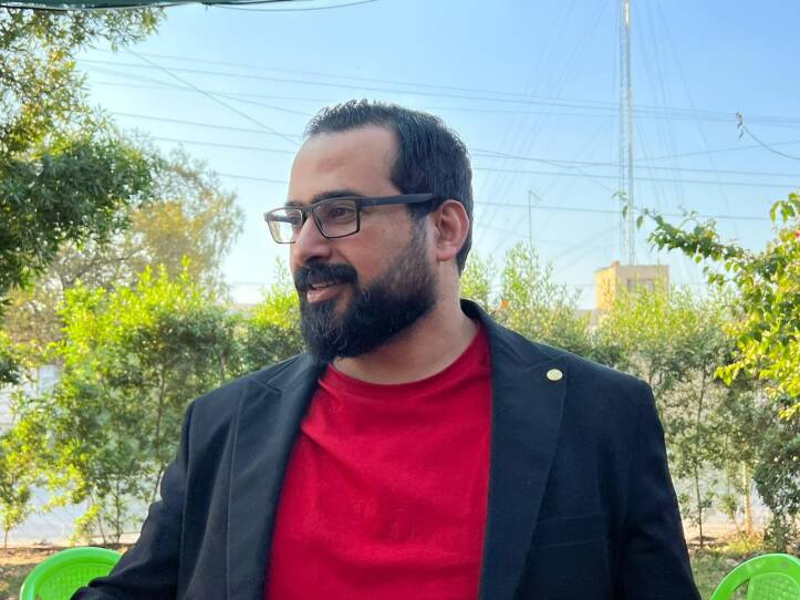 A man in a red T-shirt and black blazer looks to his right as he stands in a garden. He is surrounded by trees and standing in front of a couple plastic lawn chairs.