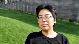A person in glasses and a T-shirt poses for the camera with a serious expression. They're standing in front of a grassy hill.