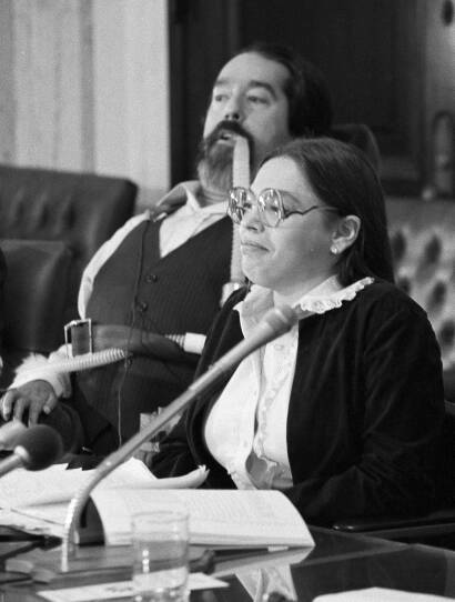 A black and white photo. Judy Heumann as a young woman sits at a table with a microphone. Behind her is Ed Roberts in a wheelchair.