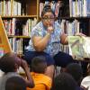 A women sits in a chair in a library full of books, her finger to her mouth, reading aloud from an illustrated book to a diverse group of children, who are sitting on the floor.