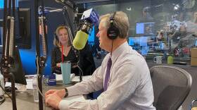 The governor sits in the radio studio with headphones on, perched in front of a microphone. Margery is in the background looking at him as he speaks.