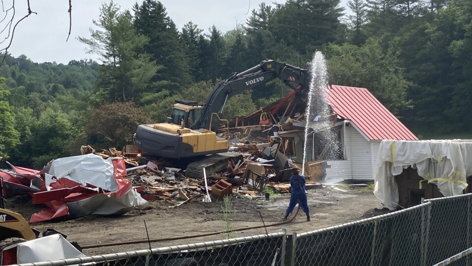 An excavator tears down a building while a person sprays a fire hose.
