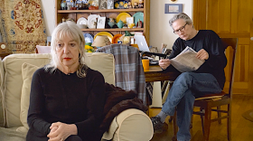 This photograph is a production still from the short film NUTS, which is about an estranged couple who are forced to live together because of the pandemic. In the foreground a woman sits in a chair. She has a concerned and contemplative expression on her face. In the background a man sits at a table, reading the newspaper.