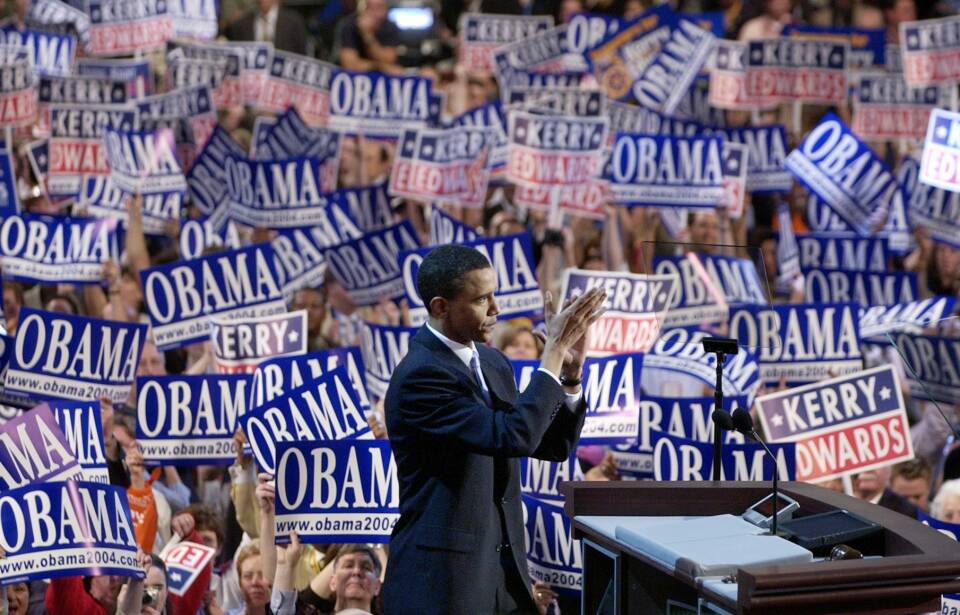 Barack Obama claps his hands together while on stage in front of a sea of political signs reading "Obama 2004" and "Kerry Edwards"