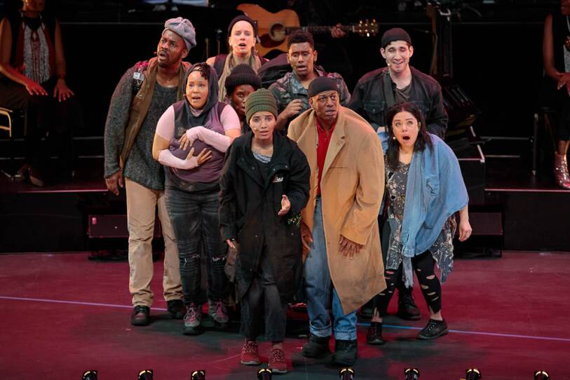 A group of actors cluster together center stage, looking off stage with anxious and fearful looks on their faces, all mid-song