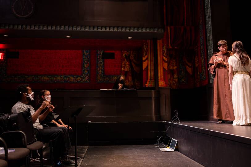 Two actors talk to each other on stage while two people wearing masks watch them and use sign language.