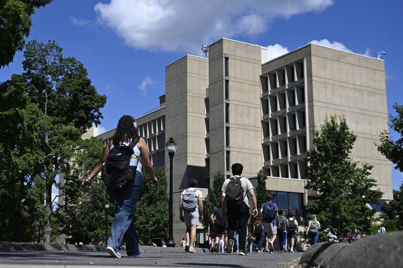 Dozens of young adults with backpacks walk down a pathway on a college campus toward a building.
