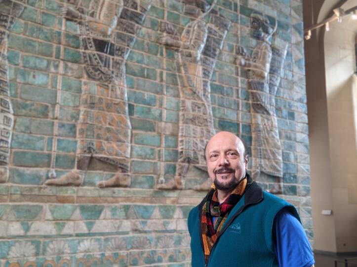 A man with a goatee smiles slightly as he stands in front of a large tile mural of Egyptian gods. He is wearing a fleece vest and plaid scarf.