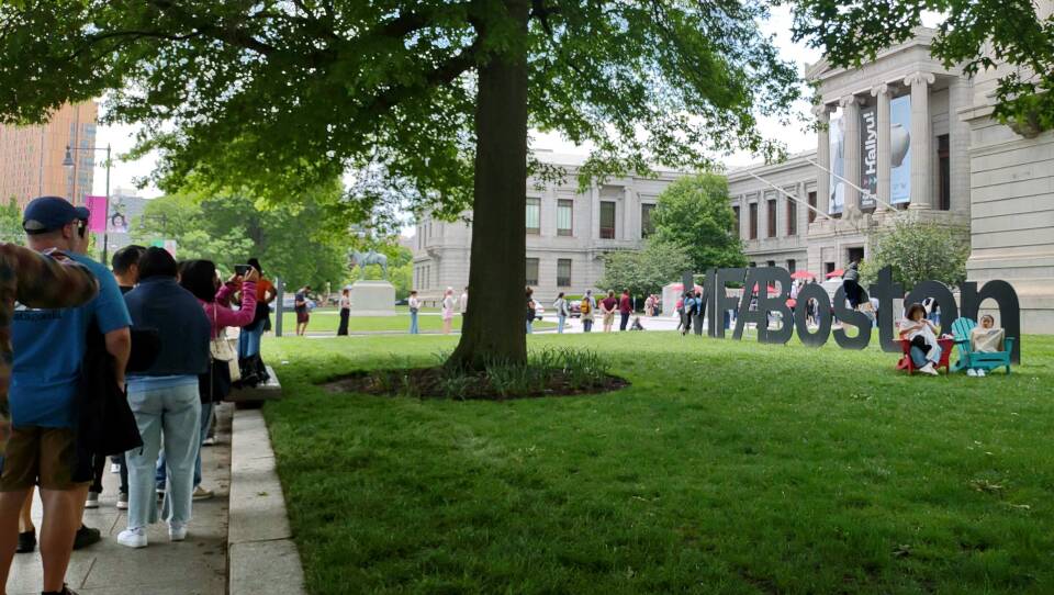 A line of people stand on the sidewalk around a large museum while others sit in chairs and pose by the MFA Boston sign in the lawn.