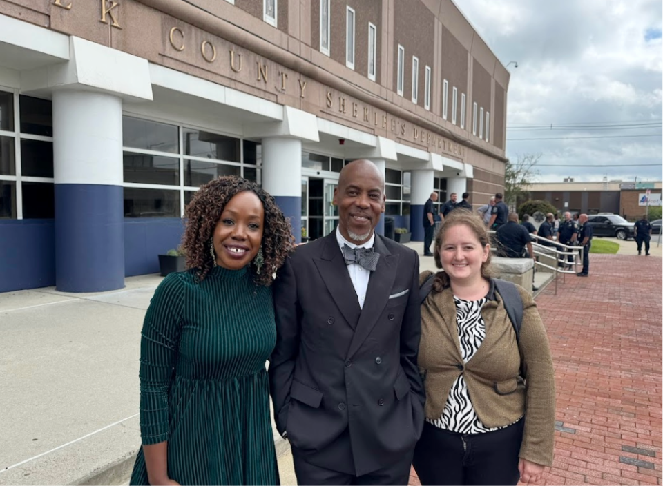 Two women and a man pose for a photo in front of the Suffolk County Sheriff's Department with a cluster of uniformed officers sitting and relaxing in the background.