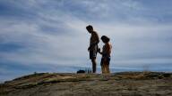 Two men in swim trunks stand atop a rocky cliff with the sun on their backs and shadows cast across their bodies.