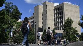Dozens of young adults with backpacks walk down a pathway on a college campus toward a building.