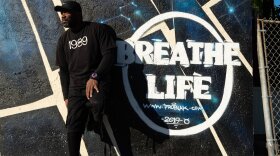In this photograph a man is outside, leaning against a wall  that has the words "Breath Life" stenciled on it I white. The words are painted  against graffiti art, which is a pattern of black and blue geometric shapes.