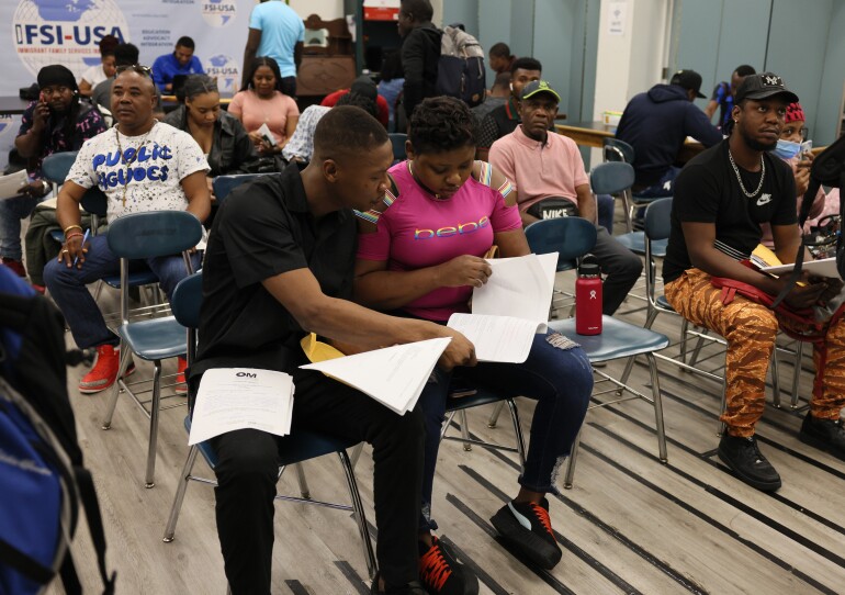 Two people fill out forms together in a crowded room of desk chairs.