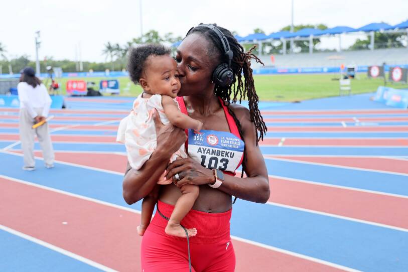 A runner wearing a numbered bib holds and kisses her baby while on an outdoor track.