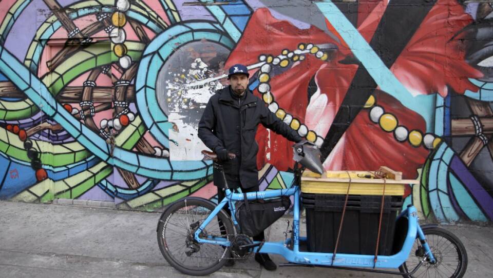 A man holding a blue bike stands in front of a colorful mural.