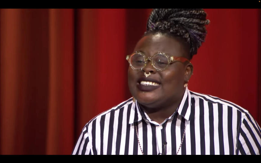 This is a headshot of poet Porsha Olayiwola. She is sitting against a red backdrop. She is wearing a black and white striped shirt. Her hair is pulled into a topknot. She is wearing round framed glasses. The rims are gold, the temples are red. She is smiling,