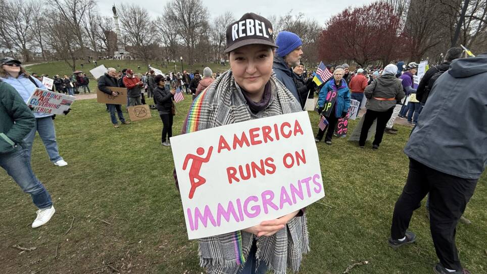 A protester in a park holds a sign that mimics the Dunkin colors and says "America runs on immigrants."