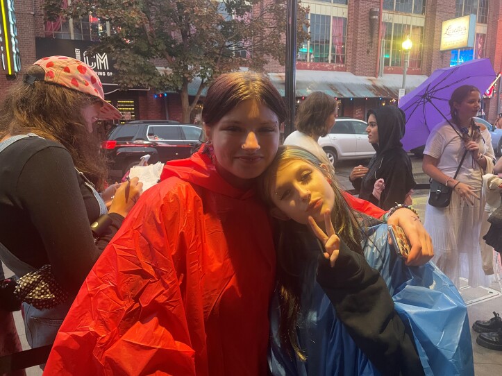 Two young women are standing out in the rain, lining up for the boygenius concert. The young woman on the left is wearing a red rain poncho. she has her arm around the young woman who is on the right. She is wearing a light blue poncho and is signaling the peace sign.