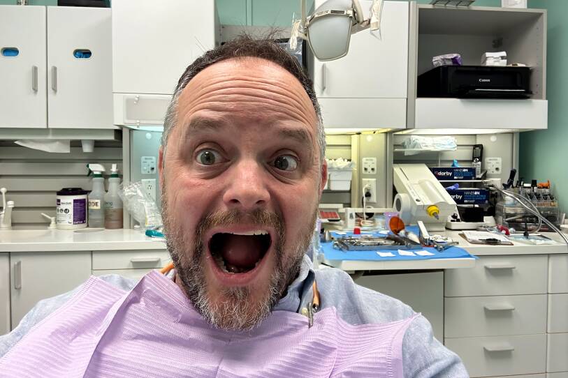 A man wearing a paper bib opens his mouth wide as he sits in a dental exam room.