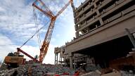 A crane labeled "JDC" operates behind a pile of rubble at a parking garage site.