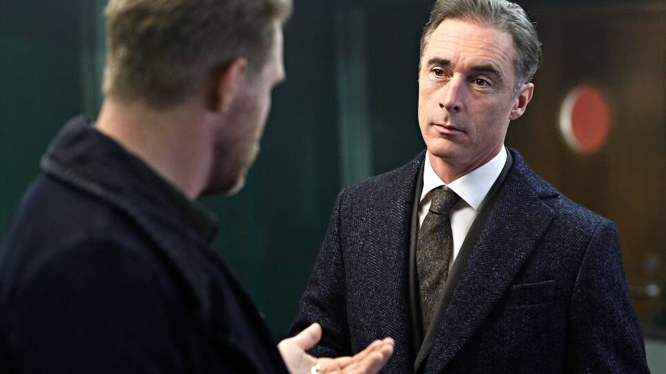 A man stands with his back to the camera. His hand is held out, with a small necklace in it. A man in a suit faces him, looking serious.