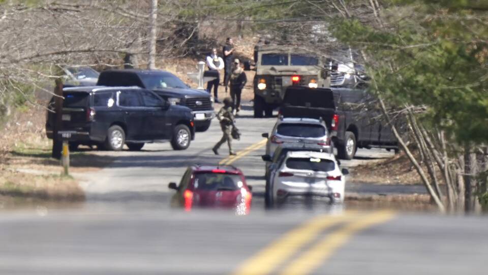 Military and civilian vehicles are clustered on a road with a few people in tactical gear walking around. It gives the impression of a busy scene.