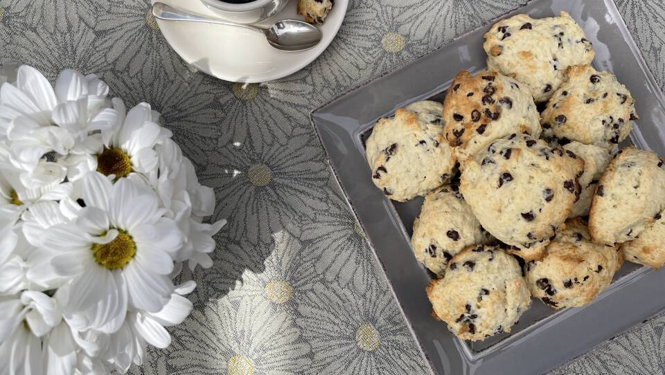 Table with a cup of coffee, a vase of white flowers, and a square plate filled with mounded, cakey chocolate chip cookies