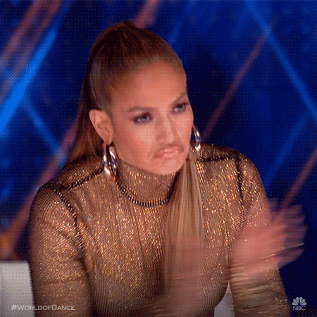 Jennifer Lopez, looking stunning in a crystal-covered dress, stands from her chair, clapping. She frowns, but it's an expression of approval.