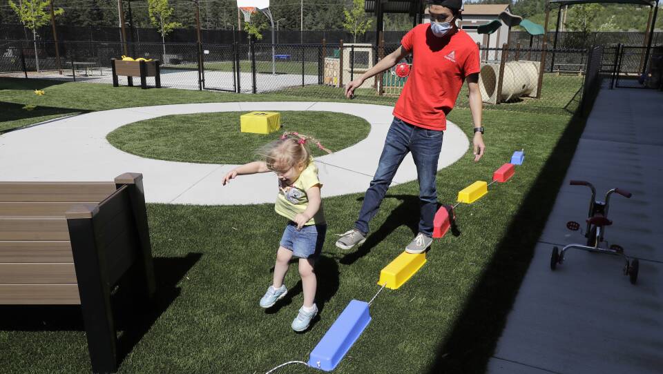 A young girl with braided hair jumps off a small, rectangular block on a grassy playground. A man behind her balances on one foot on another block.