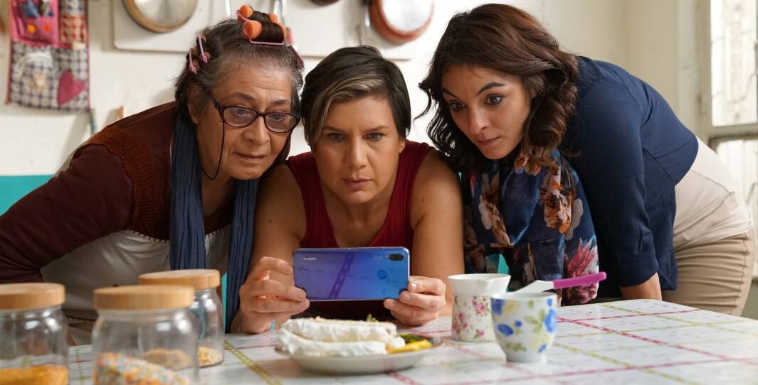 This is a scene from the film Salma's Home. Three women, who are in a kitchen , gather around a cell phone, which the woman in the middle of the trio is holding. To the left of the woman holding the cell phone is an older woman--she's wearing glasses and her hair is in curlers. The the right is a much younger woman, probably in her late 20's . All three of them have intense expressions on their faces, as though they are reading something surprising on the phone.