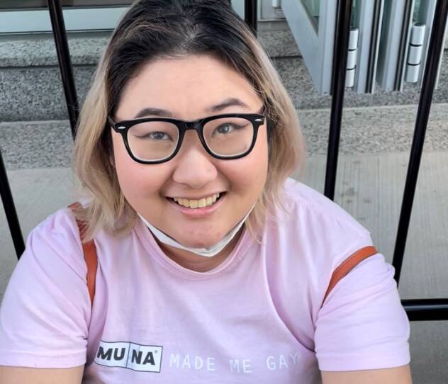 This is a headshot of a young woman. She is wearing a pink t-shirt that says "Tuna made me Gay." she is wearing glasses. her hai, which reaches her shoulders, is dark on the top, blonde on the bottom. She's smiling.