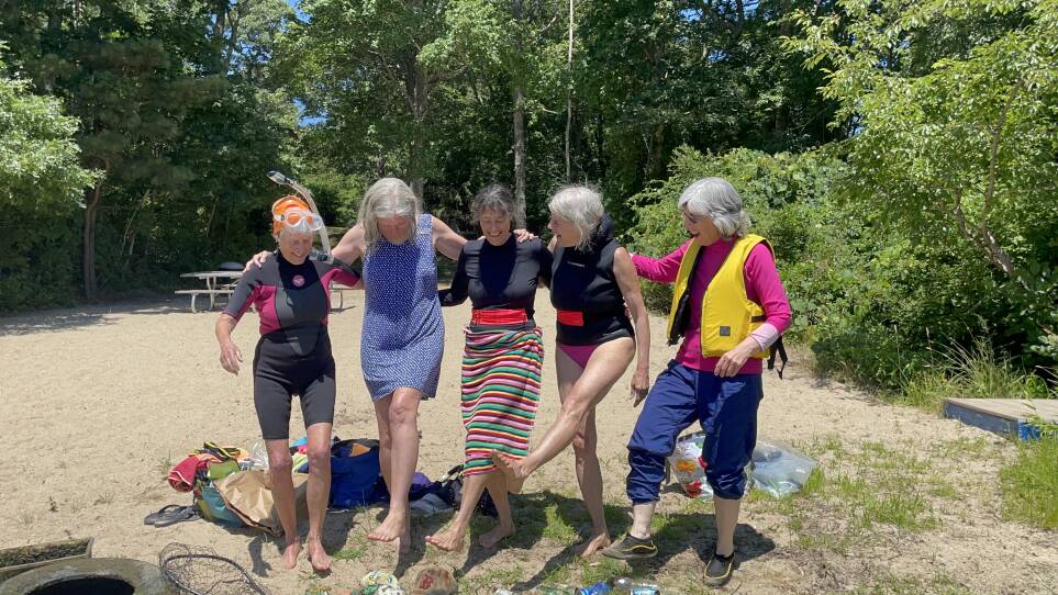 Five older women in swimwear and life vests, with their arms on one another's backs, each kick out one leg as they dance on the sand.